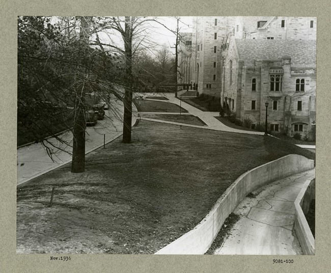 Black and white photograph of pathways intersecting open green area with the occasional tree around, in front of a large stone building.