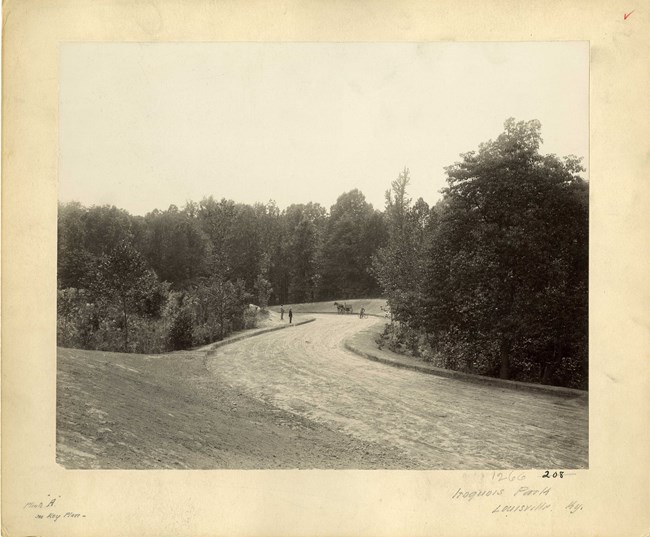 Black and white of curving dirt road around small hills with dense tree plantings. There is a horse and people on the road.