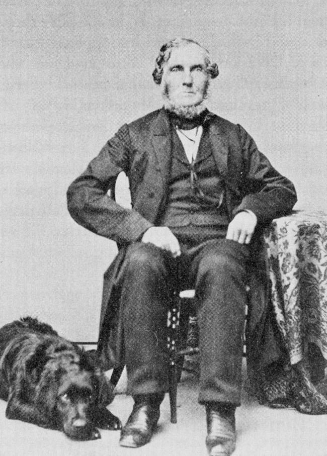 Black and white photograph of elderly man with white beard and hair seated at a chair in a suit with a black dog laying by its feet