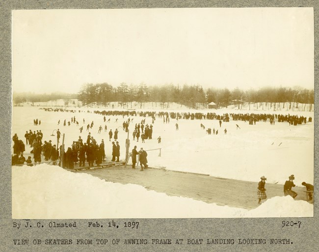 Black and white of body of water frozen solid with ice on stop and many people skating on water. Behind the water is a snowy area with trees without leaves.