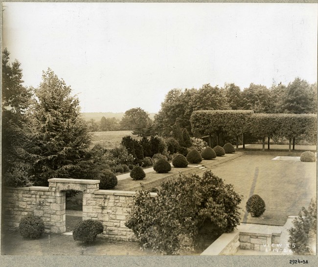 Black and white photograph of rectangular garden area enclosed by a stone wall and entrance. The edges of the garden is full of plantings, but the center is an open grassy rectangle.