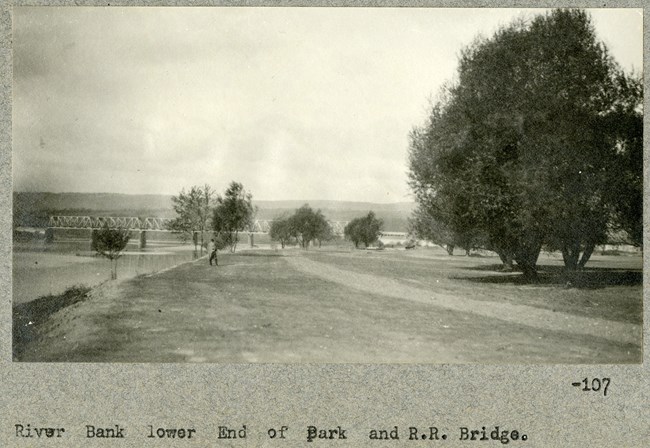 Black and white of flat grassy land by water with some trees on the edges with a bridge and mountains in the distance