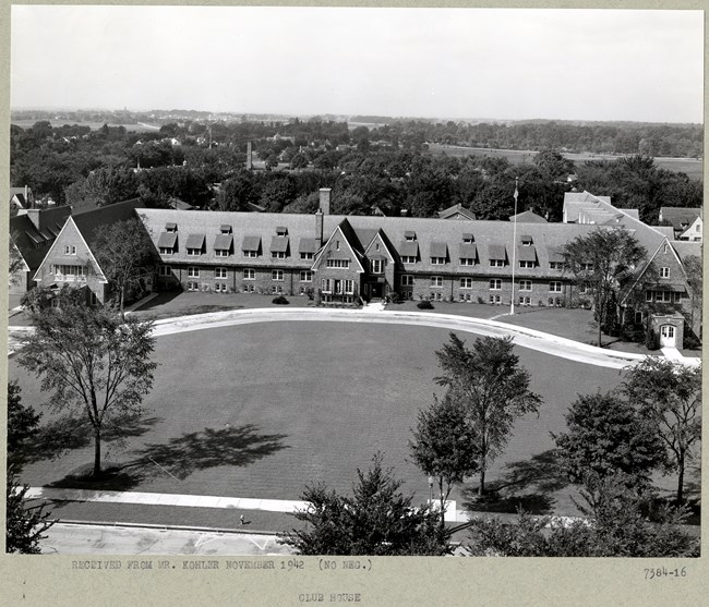 Black and white of a large building with curving road leading up to it, trees lining around it, and a large open green space in front.