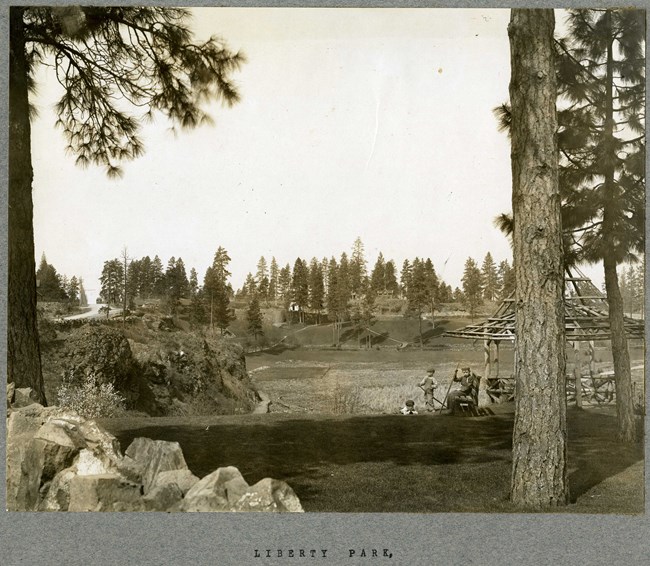 Black and white of hilly park filled with grass and trees. There is a valley in the middle of the hill and a bench with boys around it.