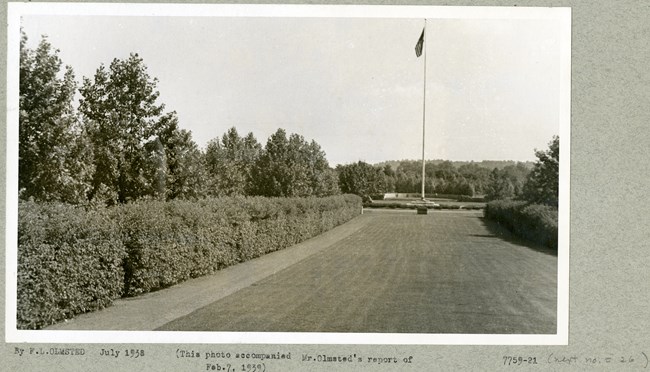 Black and white photograph of an open rectangular grassy area with a row of bushes on the long sides, followed by trees. At the end of the grassy area is an American flag and pole