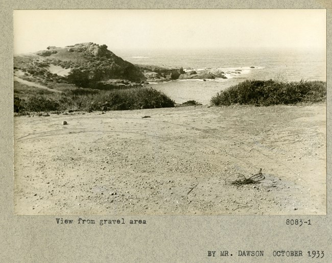 Black and white photograph of gravel area overlooking hills covered with shrubs jutting out into the ocean