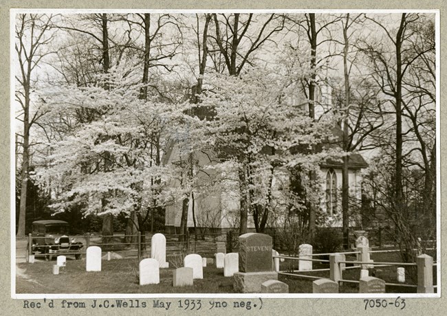 Black and white photograph of group of gravestones, with stand of trees with light leaves surrounding a building.