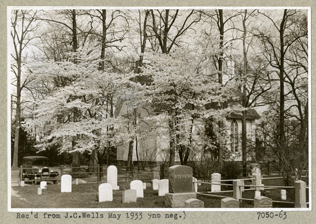 Black and white photograph of group of gravestones, with stand of trees with light leaves surrounding a building.