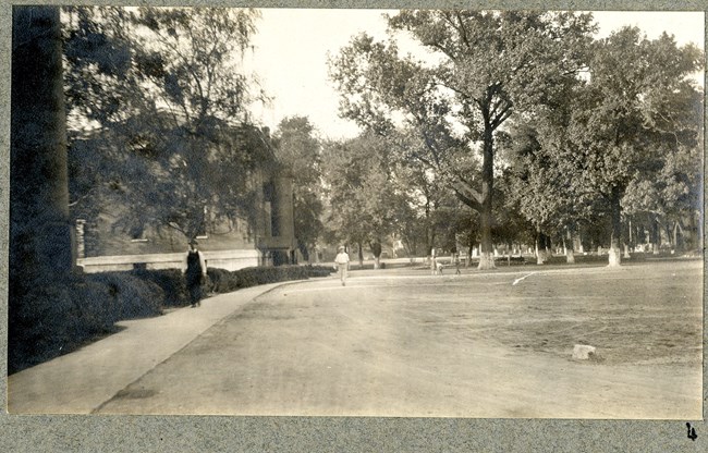 Black and white of dirt area with path going around and trees and shrubs on the edge and in the distance. People walk around the path.