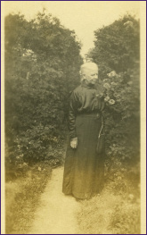 Black and white photograph of blonde woman standing on dirt path wearing a black dress. On both sides of the path are large bushes, taller than her
