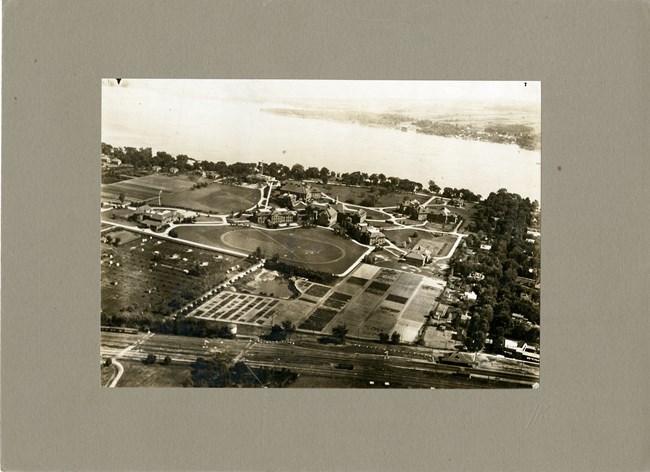 Black and white aerial photograph of college campus with lots of green space, curved lines, and buildings. In the distance is a large body of water.