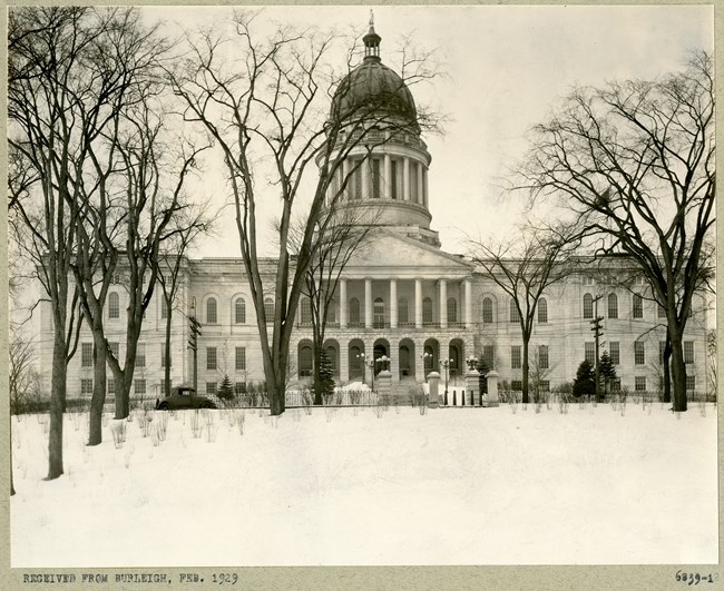 Black and white of large white building with dome with flat area in front covered with grass, some trees and shrubs.