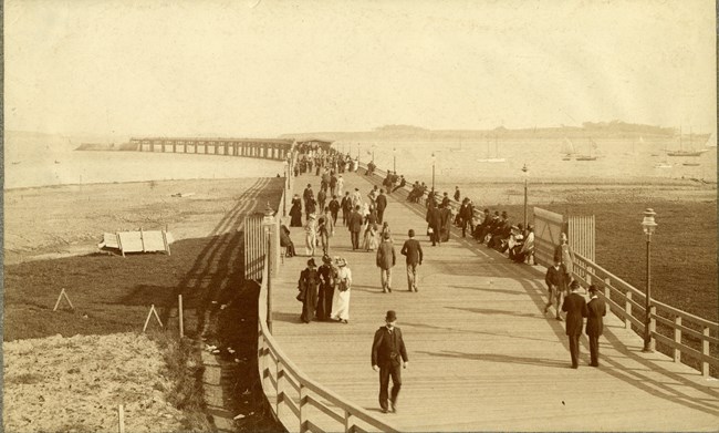 black and white of long wooden dock going into the water with many people walking on dock, with sailboats in the water.