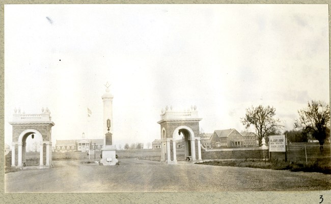 Black and white photograph of two stone arches leading to large open green space with some buildings in the distance