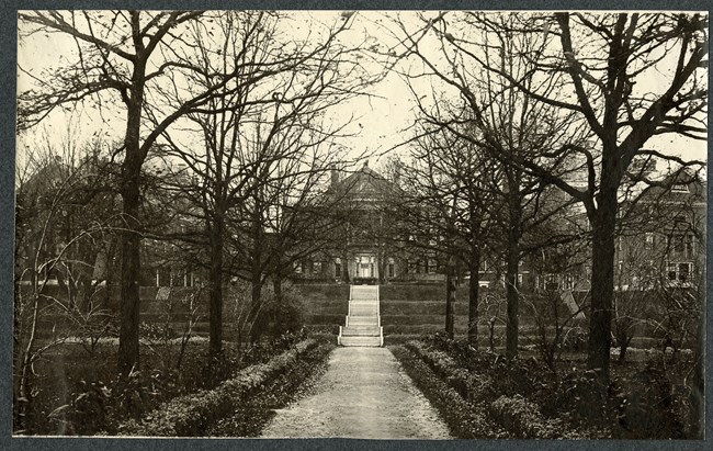 Black and white photograph of path leading up to large building. The path is lined with trees and there are stairs up to the building. The whole image is symmetrical.