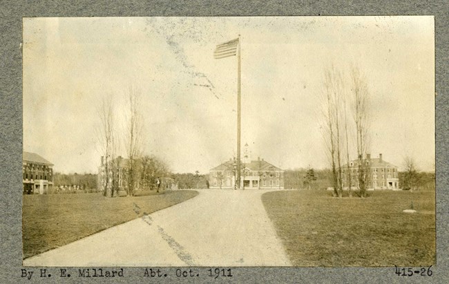 Black and white photograph of long dirt road leading to a circular area with a flagpole at the center and buildings all around the circle, with few trees on the grassy area