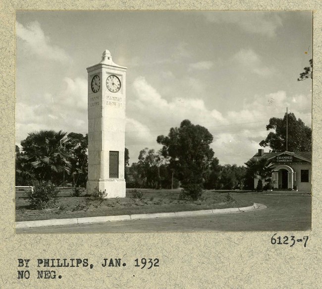 Black and white photograph of large white tower with clock at the top center in a grassy area surrounded by few trees.