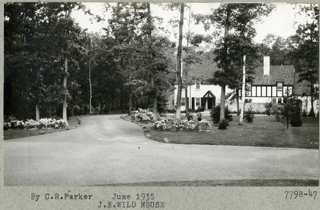 Black and white of dirt road lined with trees and flat grassy area with a house at the end of the road.