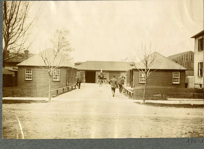 Black and white of dirt road with a small house on both sides and one at the end, with people walking down the street.