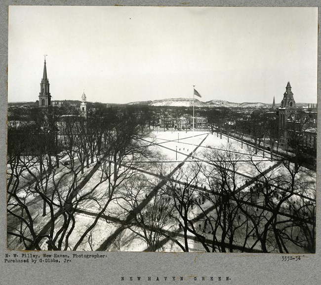 Black and white aerial of large open green space with paths cutting through, with buildings on its edges and mountains in the distance