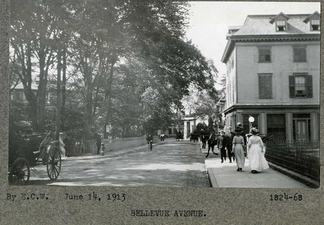 Black and white of street with houses on one side, trees and green space on the other, with many people and carriages going down the road.