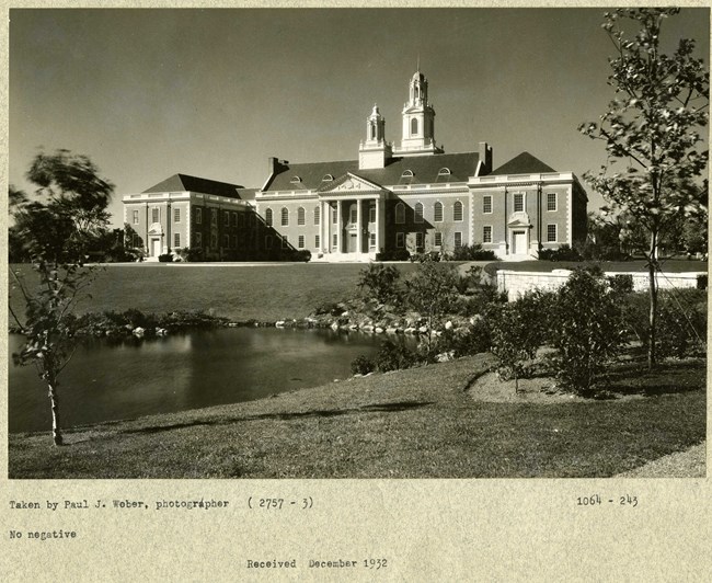 Black and white photograph of large brick building with two towers rising from the top. There is an open green space in front of the building, with a body of water in front of that
