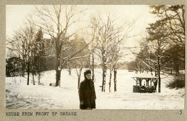 Black and white of person standing on snow with trees in background without leaves, and a hill leading to a house. There is also a gazebo on the snowy hill area.