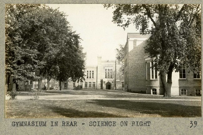 Black and white photograph of brick building, like a castle, in front of an open green space with scattered trees and a walking path cutting through it
