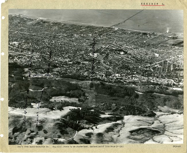 Black and white aerial of community with many homes together. On one side is a hilly area, on the other is a flat area.