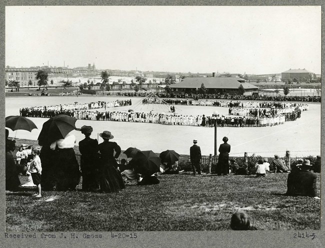 Black and white of large flat open space with grassy hill around it with many people sitting on the hill. On the flat area, many people form a rectangle with a pole in the middle.