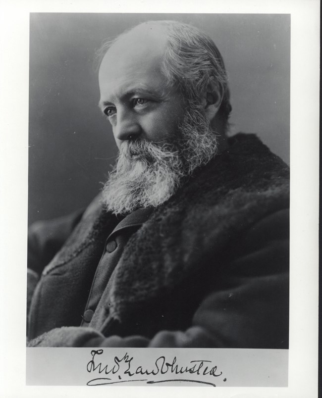 Black and white photograph of man with thick white beard and a bald top of the head, with some hair in the back, posing for a picture in a jacket.