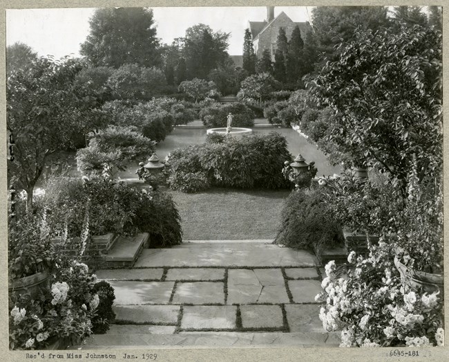 Black and white photograph of rectangular garden with fountain statue in the center. There are stone stairs leading down to the area, and that is surrounded by plants and shrubs
