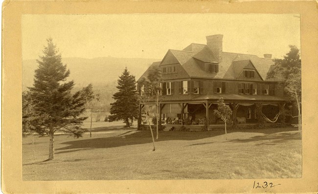 Black and white photograph of large home with many windows and a porch wrapping around the home, with a hammock on one side. The home is on a large grassy area, with a few scattered trees. In the distance is a large wooded mountain