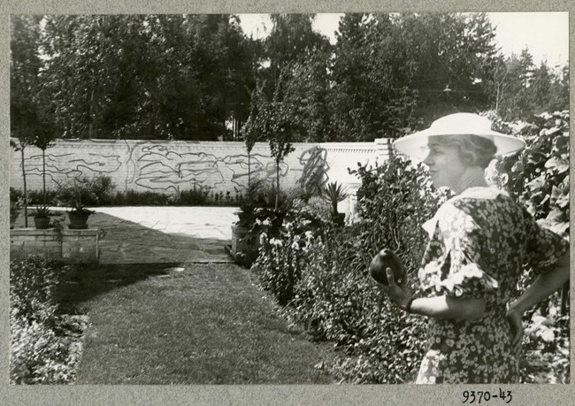 Black and white of flat grassy area with plantings on both side and a wall in the distance with a woman walking along the path.