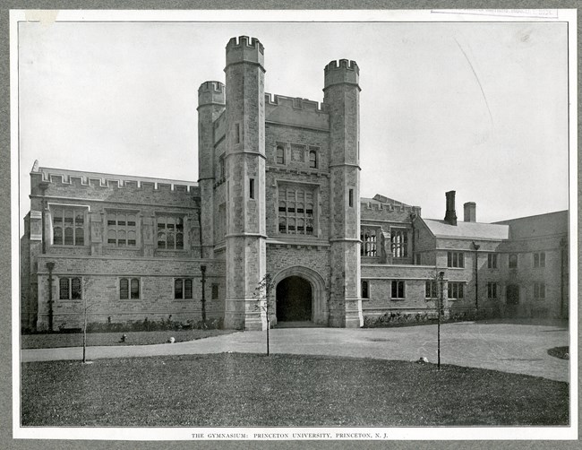 Black and white of flat grassy area with paths leading up to a large building, which is lined by shrubs.