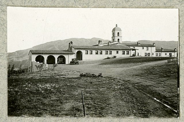 Black and white of large white building with tiled roof on hill with mountain rising in distance.