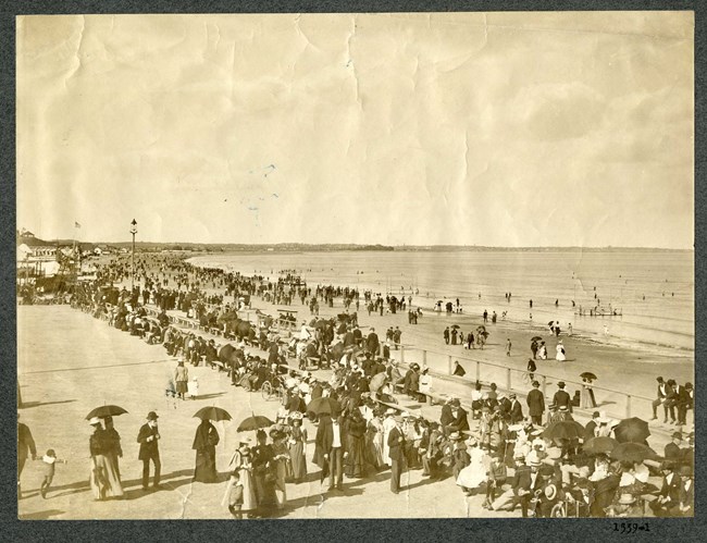 Black and white photograph of large crowd of people gathered on shore of beach
