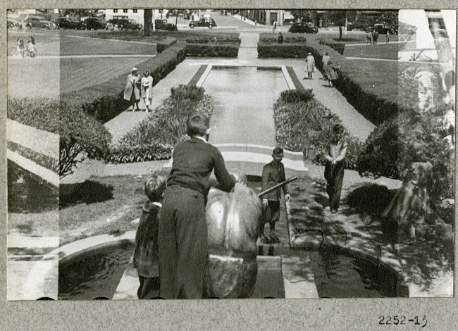 Black and white of children playing on statue of squatting man overlooking square body of water lined with shrubs, with grass on the other side.