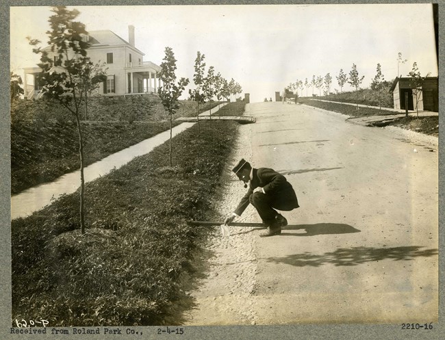 Man in suit and hat smoking pipe bends down on side of dirt road that leads uphill with trees following it.