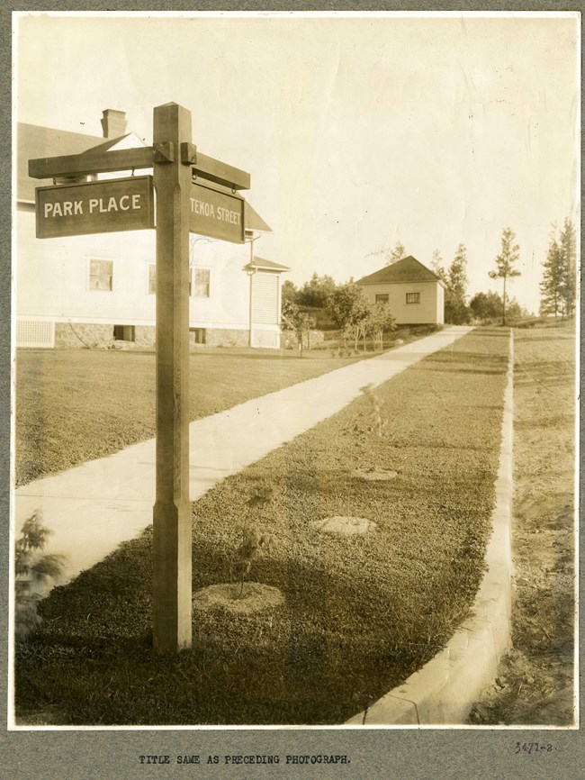 Black and white photograph of sidewalk with grass on both sides and a street on one side and a few buildings on the other. At the end is a street sign pointing to Park Place