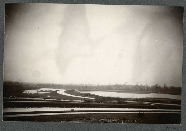 Black and white of flat land with body of water and roads going around it, with many grassy areas.