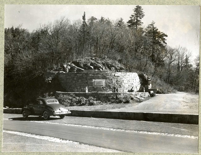 Black and white of stone spiral structure on hill wiht trees in background and stairs leading up stones.