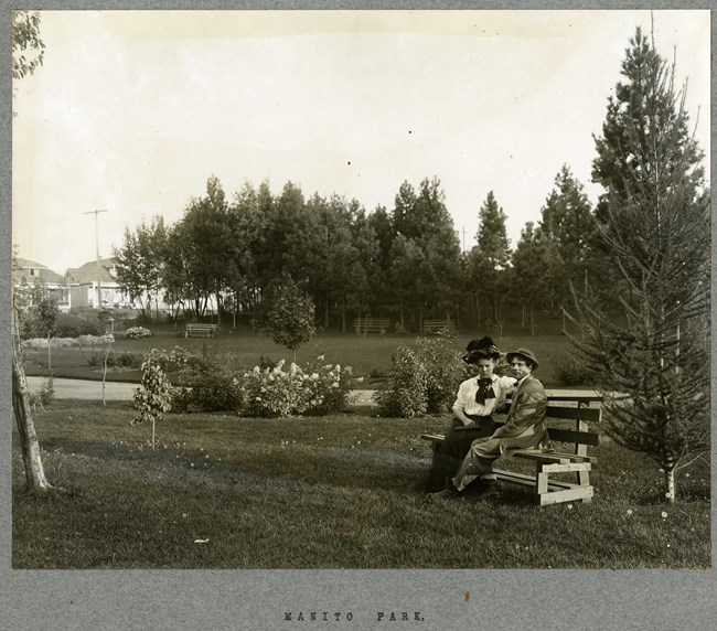 Black and white of flat grassy area with dirt path and flowers in the back. Two women sit on a bench looking at the camera