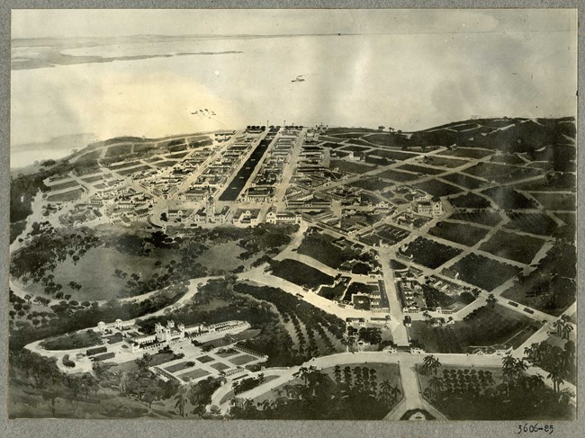 Black and white photograph of birds-eye view of a city by the water dominated by green patches between the buildings