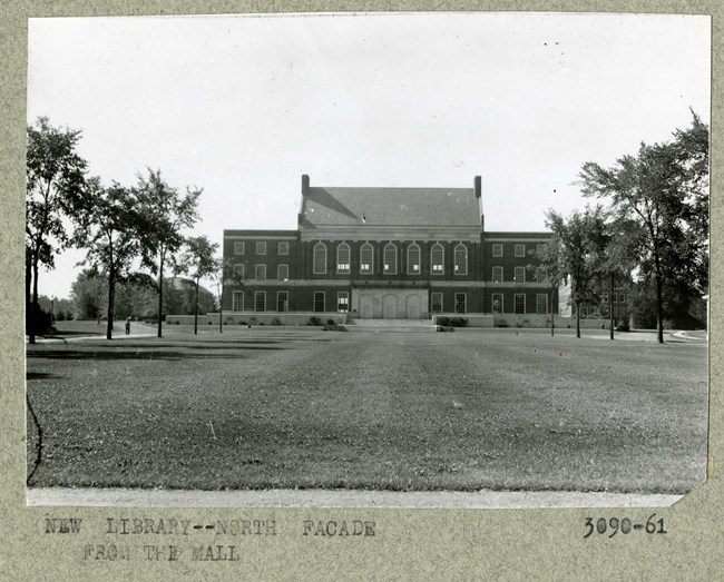 Black and white of flat grassy area with trees on the edges with a large brick building at the end of the grass.