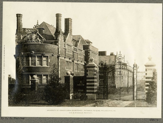 Black and white of large building with gate in front and road leading into it with a flat grassy area outside with some shrubs on the fence.