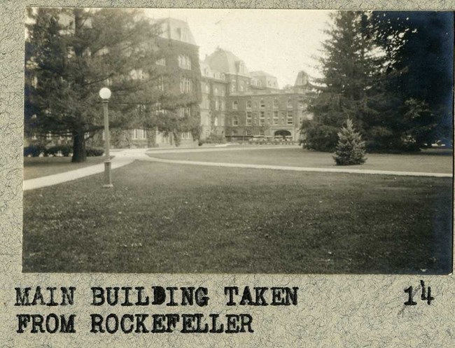 Black and white of flat grassy area with paths cutting through, and buildings in the background.