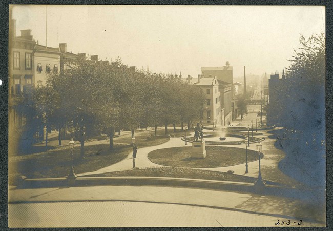 Black and white of park between two roads with homes on the other side, with trees and grass lining by the roads, and circular green spaces on the cement paths with a fountain at one circle.