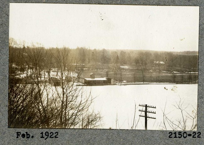 black and white of large body of water covered with snow with building on edge and grassy area behind with trees, though they don't have leaves.