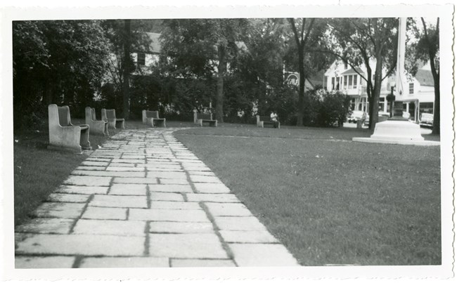 Black and white of curving stone path around grassy area with benches on one side, a flagpole in the middle, and buildings in the distance